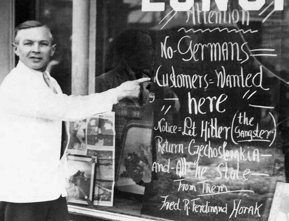 <i>Chủ nhà hàng người Mỹ Fred Horak, người gốc Séc ở Praha, bên cửa sổ nhà hàng của mình. Dòng chữ: "Khách người Đức không được chào đón ở đây. Hãy để Hitler trả lại Tiệp Khắc và tất cả những gì mà ông ta đã cướp đi từ họ".</i>