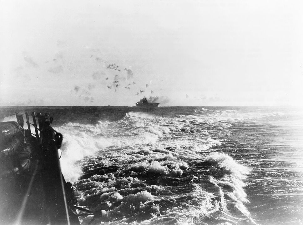 Hình ảnh Enterprise được nhìn thấy từ một tàu hộ tống trong lúc bị máy bay ném bom bổ nhào của Nhật Bản tấn công. Một đám cháy dữ dội đang bùng phát ở bệ pháo 5 inch phía sau mạn phải của tàu, do một quả bom đánh trúng làm bốc cháy đạn dược dự trữ. Chú ý mức độ hoả lực phòng không dày đặc từ tàu sân bay. (NHHC)