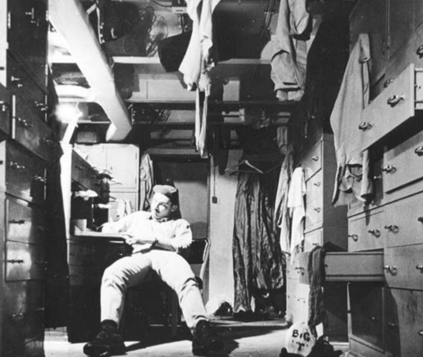 Một sĩ quan đang viết thư trong không gian chật hẹp của khu khoang ngủ dành cho sĩ quan cấp dưới. Mặc dù không chật như trên tàu ngầm, nhưng điều kiện sinh hoạt trên tàu sân bay vẫn đông đúc đối với mọi cấp bậc, và mọi khoảng trống có thể sử dụng đều được tận dụng. (Lưu trữ Quốc gia)