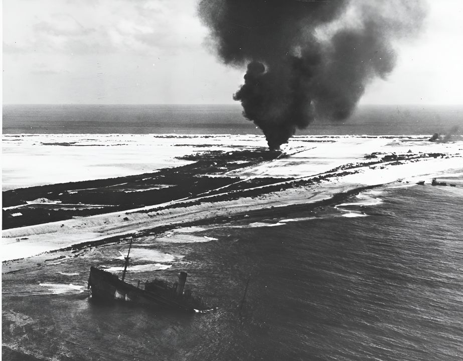 Ảnh chụp từ trên không, góc nhìn xiên thấp của Đảo Wake vào thời điểm cao trào của cuộc tấn công bằng máy bay từ tàu sân bay và pháo kích của tàu chiến Mỹ. Một đám cháy bùng lên gần sân bay, trong khi ở phía trước là xác một con tàu Nhật Bản bị mắc cạn sau khi bị Thủy quân Lục chiến bảo vệ căn cứ tiêu diệt vào tháng 12 năm 1941. (NHHC)