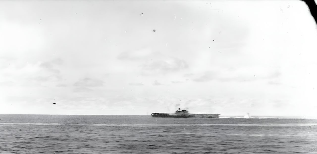 Chụp từ một tàu hộ tống, bức ảnh nổi bật này cho thấy Yorktown đang bị tấn công bằng bom bổ nhào. Một máy bay ném bom bổ nhào Type 99 của Hiryu – mất phần đuôi – sắp rơi xuống biển phía trước Yorktown. Một chiếc khác hiện ra phía trên Yorktown sau khi hoàn thành cú bổ nhào, nhưng quả bom của nó đã trượt về phía sau. (NHHC)