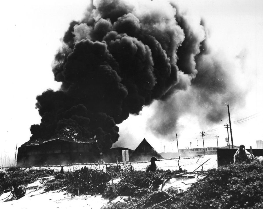 Các bồn chứa dầu đang bốc cháy trên đảo Sand, Midway, sau cuộc không kích của Nhật Bản vào sáng ngày 4 tháng 6 năm 1942. Những bồn chứa này nằm gần bờ biển phía nam của đảo Sand lúc bấy giờ. Cảnh này nhìn từ bãi biển vào sâu trong đất liền. Ba chú chim hải âu Laysan (chim hải âu Gooney) con hiện rõ ở phía trước.